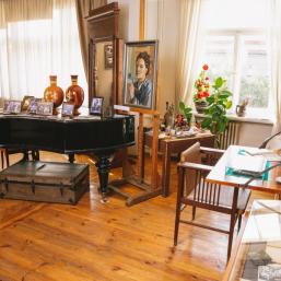 A room with a wooden floor, piano, paintings, and furniture resembling an artist’s studio or museum interior.