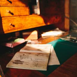 A wooden writing desk with an open sketchbook and drawings illuminated by warm sunlight.