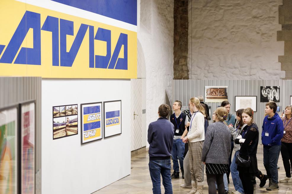 Jauniešu grupa stāv pie informatīvā tablo ar vilcienu kustības laikiem, viena meitene paceļ roku un rāda uz ekrānu.