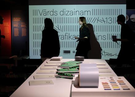 Cilvēki tumšā izstāžu telpā stāv pie galda ar dizaina materiāliem, fonā redzams liels ekrāns ar tekstu.