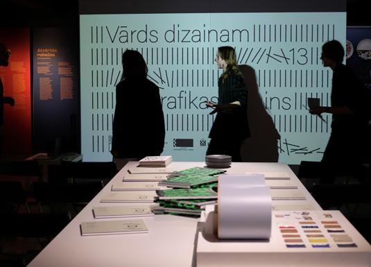Cilvēki tumšā izstāžu telpā stāv pie galda ar dizaina materiāliem, fonā redzams liels ekrāns ar tekstu.