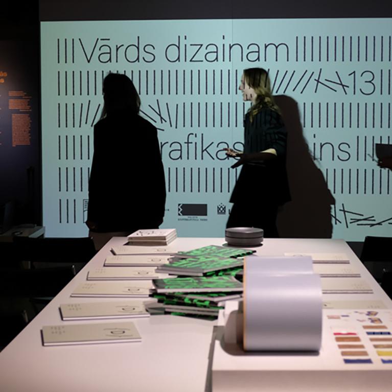 Muzeja izstāžu telpā pie balta galda stāv vairāki cilvēku silueti, fonā redzams liels ekrāns ar tekstu Vārds dizainam un grafiskiem elementiem.