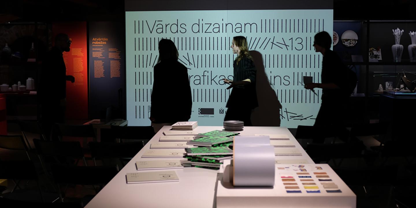 Muzeja izstāžu telpā pie balta galda stāv vairāki cilvēku silueti, fonā redzams liels ekrāns ar tekstu Vārds dizainam un grafiskiem elementiem.