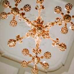 A gilded historic chandelier with ornate decorations on a white ceiling panel.