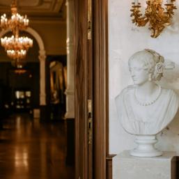 A white marble bust of a woman by the wall in an elegant museum interior with chandeliers and display cases in the background.
