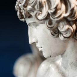 Close-up of a marble sculpture showing the profile of a young man with curly hair against a blue background.