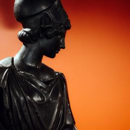Dark bronze sculpture of a female figure wearing a classical robe and helmet, set against a red background.