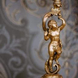 Zeltīta dekoratīva lustra ar putti figūru un ornamentāliem rotājumiem uz rakstaina fona.