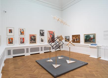 Izstādes zāle ar 20. gadsimta mākslas darbiem — gleznām, fotomontāžām un konstruktīvisma iedvesmotām telpiskām instalācijām, eksponētām uz baltām sienām un koka grīdas fona.