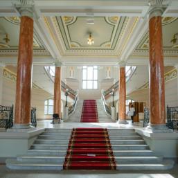 The grand vestibule of the Latvian National Museum of Art with marble stairs, a red carpet, and decorative columns.