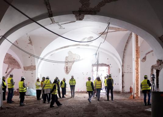 Cilvēku grupa ar aizsargķiverēm un vestēm aplūko plašu vēsturisku zāli būvniecības laikā.