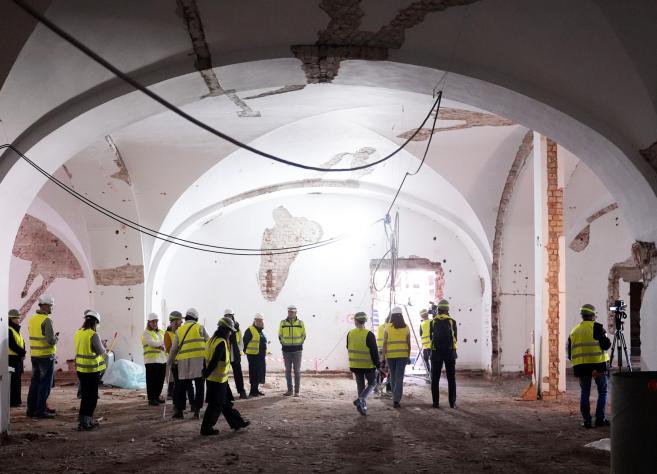 Cilvēku grupa ar aizsargķiverēm un vestēm aplūko plašu vēsturisku zāli būvniecības laikā.