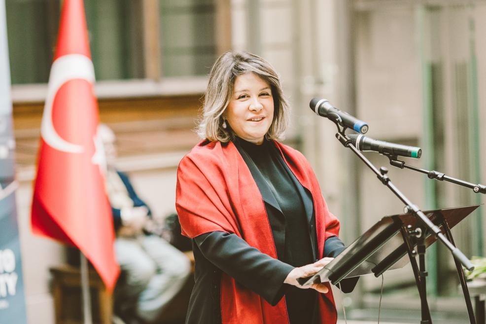A woman with a red shawl gives a speech at the microphone with a flag in the background.