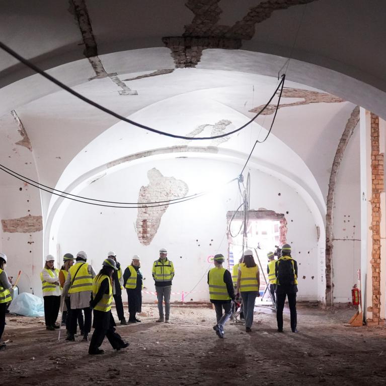Group of people wearing helmets and vests explore a large historic hall during construction.