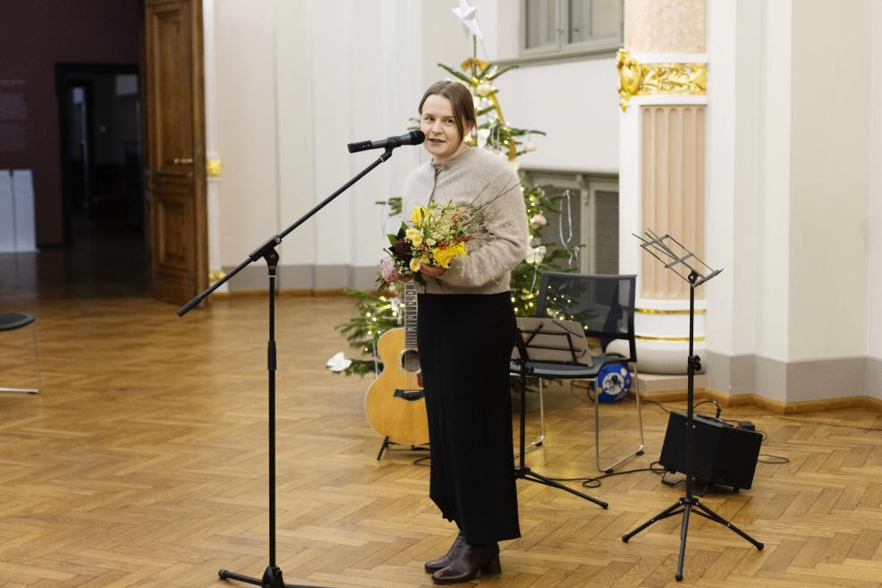 Sieviete ar ziedu pušķi runā pie mikrofona svinīgā pasākumā muzeja zālē, fonā redzama Ziemassvētku egle un mūzikas instrumenti.
