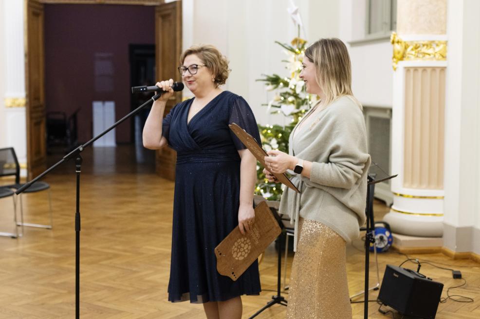 Divas sievietes stāv muzeja zālē pie mikrofona svinīgā pasākumā, viena runā, bet otra tur plāksni, fonā redzama Ziemassvētku egle un mūzikas aprīkojums.