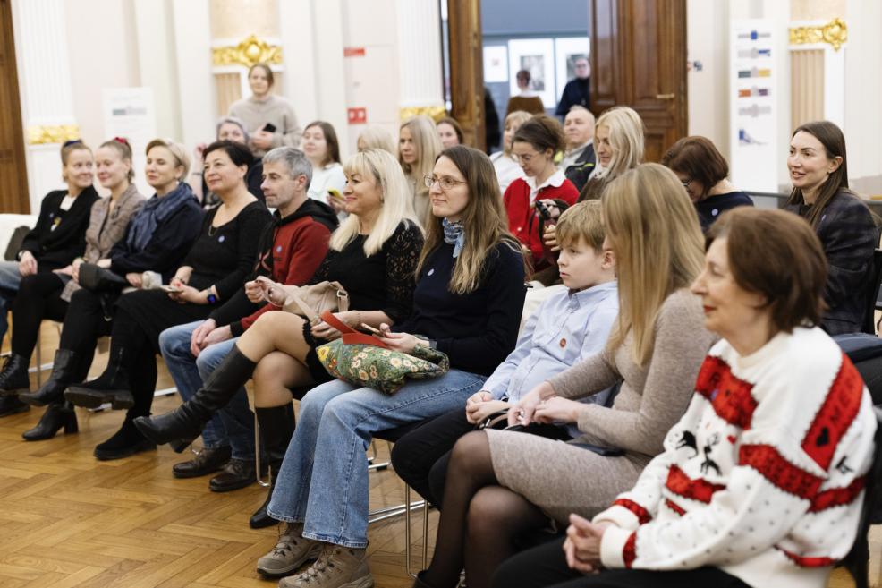 Pasākuma apmeklētāji, tostarp pieaugušie un bērns, sēž rindās muzeja zālē un klausās norisi svinīgā, draudzīgā atmosfērā.