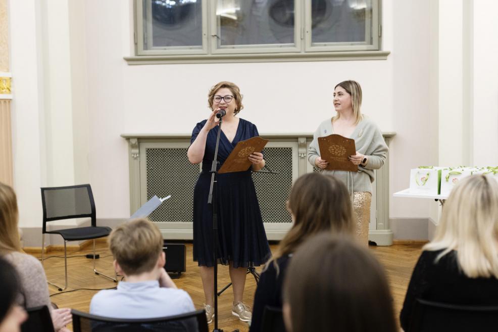 Divas pasākuma vadītājas uz skatuves muzeja zālē uzrunā auditoriju ar mikrofonu un planšetēm rokās, skatītāji sēž priekšplānā.
