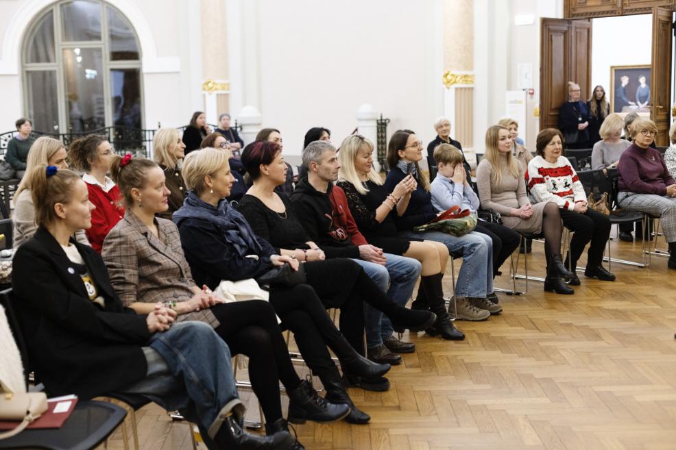 Pasākuma apmeklētāji sēž rindās muzeja zālē un uzmanīgi klausās notiekošajā, dažiem klēpī ir personīgās mantas un piezīmes.