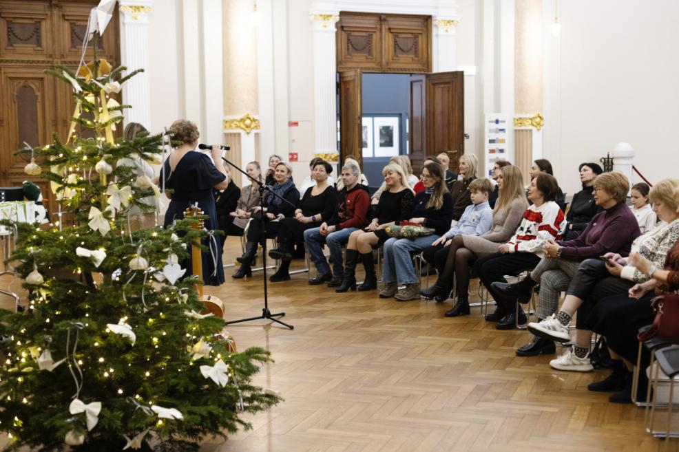 Ziemassvētku eglītes priekšplānā un auditorijas priekšā muzeja zālē pie mikrofona uzstājas runātāja, kamēr klausītāji sēž rindās un seko notikumam.
