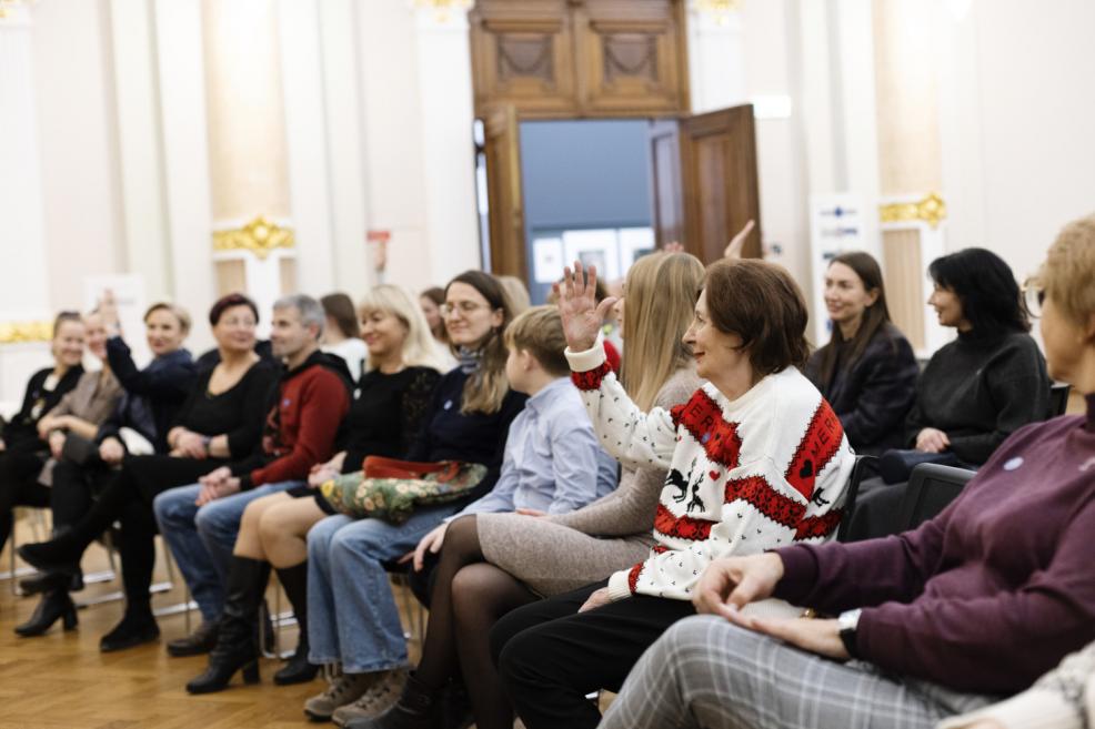 Muzeja zālē sēdoša auditorija pasākuma laikā, kur viena no klausītājām paceļ roku, iesaistoties sarunā, kamēr pārējie uzmanīgi klausās.