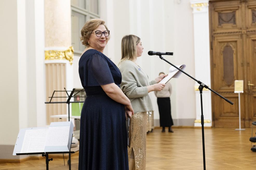 Divas pasākuma vadītājas muzeja zālē pie mikrofona, viena stāv smaidot pret skatītājiem, kamēr otra lasa tekstu no lapas.