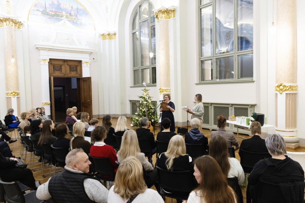 Plašā muzeja zālē klausītāju pilna auditorija sēž rindās un klausās divas pasākuma vadītājas pie mikrofona, pie svētku egles.