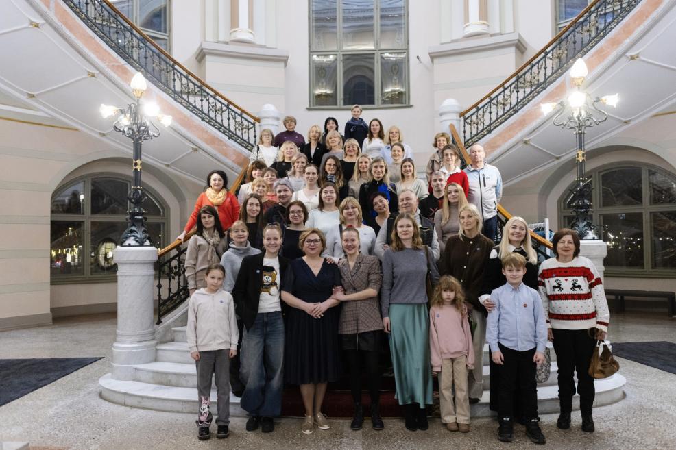 Plaša dalībnieku grupa, tostarp pieaugušie un bērni, pozē kopbildei uz muzeja centrālajām kāpnēm zem dekoratīvām lampām un balkoniem.