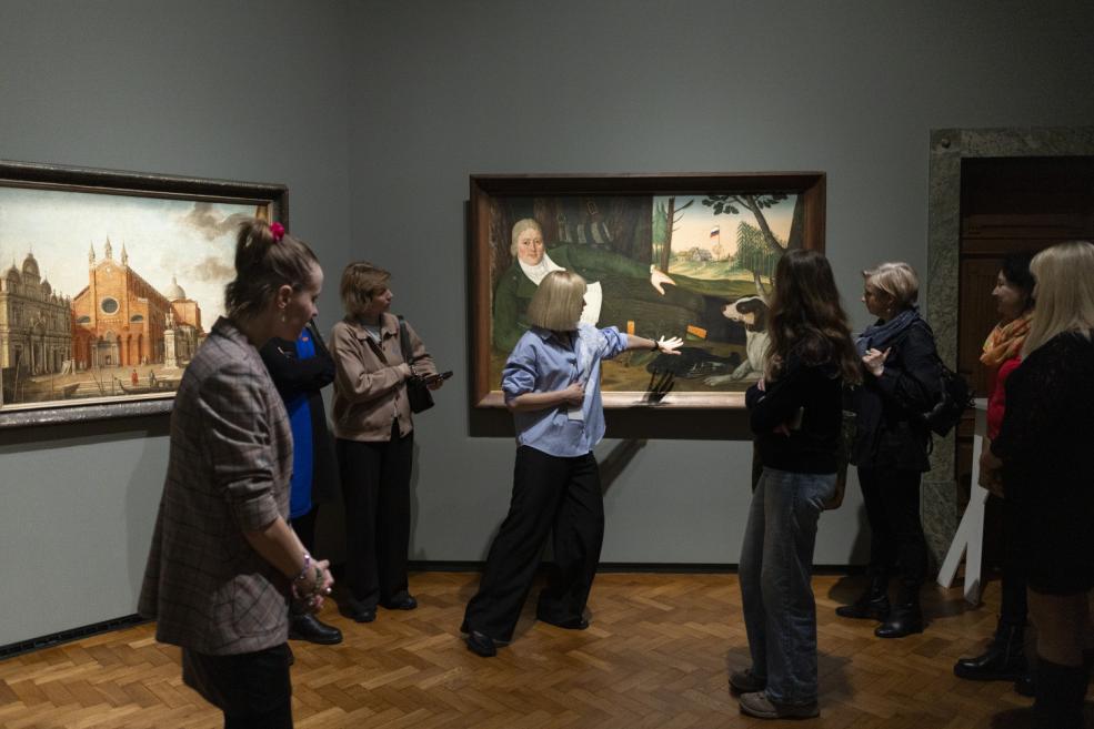 Grupa apmeklētāju muzeja zālē klausās gida skaidrojumu, kamēr viņa ar roku norāda uz gleznas detaļām pie sienas.