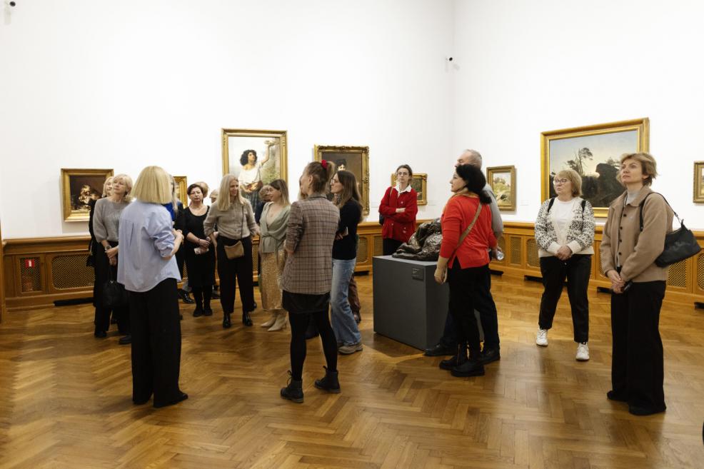 Apmeklētāju grupa muzeja zālē stāv puslokā ap gidu, klausoties stāstījumu par izstādītajiem mākslas darbiem pie sienām.