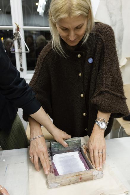 Darbnīcā sieviete kopā ar citu dalībnieku strādā ar sietspiedes rāmi, ar rokām izlīdzinot krāsu uz papīra.