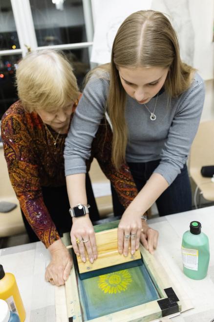 Divas sievietes darbnīcā kopīgi strādā ar sietspiedes rāmi, ar spiedpadevi uzklājot krāsu uz auduma ar zieda motīvu.