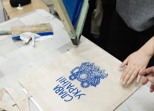 Darbnīcas laikā uz auduma somas tiek drukāts zils grafisks raksts ar uzrakstu “Slava Ukrainai!”, redzamas rokas un sietspiedes aprīkojums.