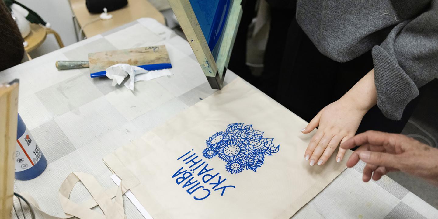 Darbnīcas laikā uz auduma somas tiek drukāts zils grafisks raksts ar uzrakstu “Slava Ukrainai!”, redzamas rokas un sietspiedes aprīkojums.