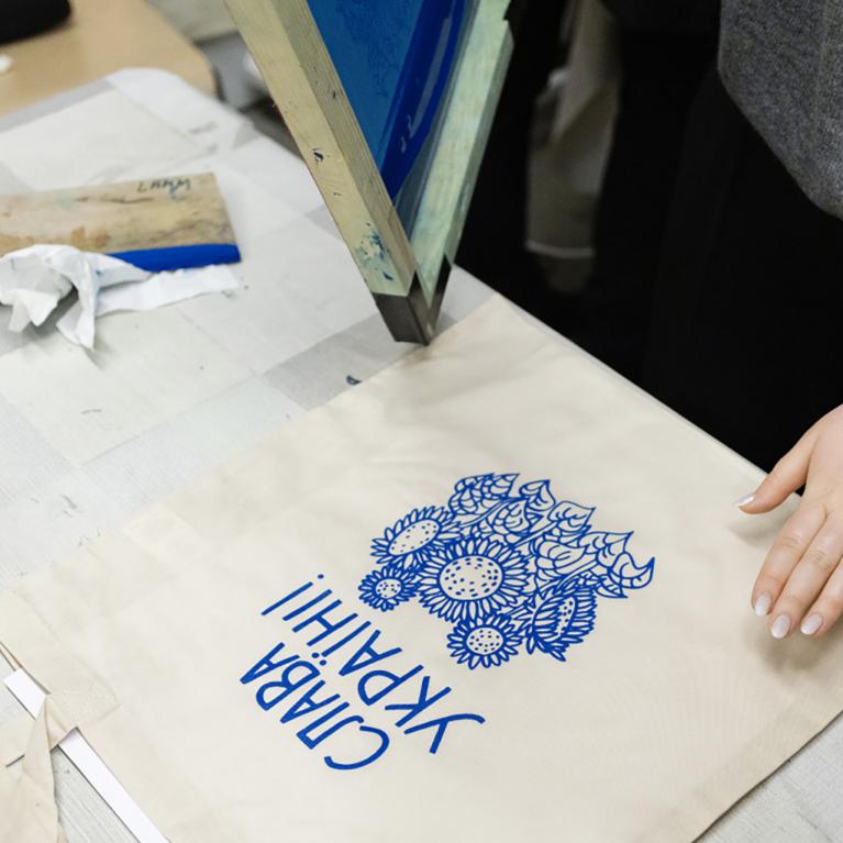 Darbnīcas laikā uz auduma somas tiek drukāts zils grafisks raksts ar uzrakstu “Slava Ukrainai!”, redzamas rokas un sietspiedes aprīkojums.