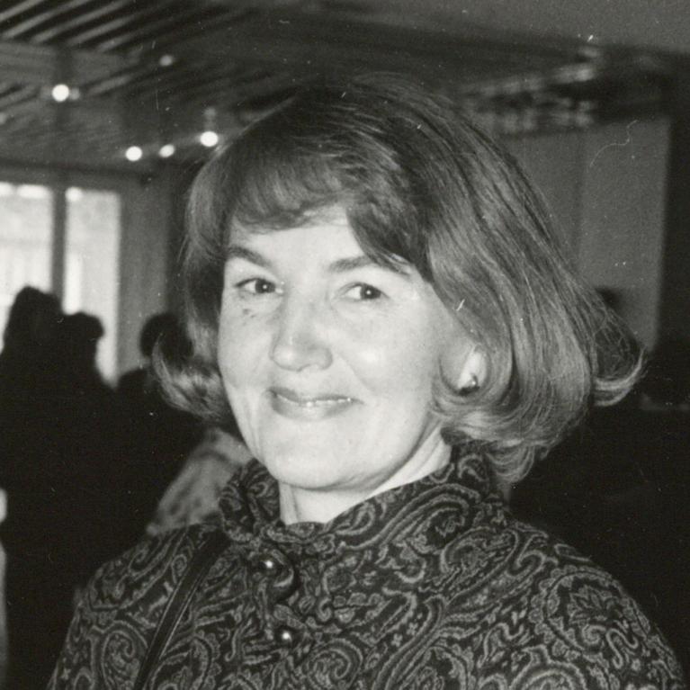 Melnbalta fotogrāfija ar sievieti pusportretā, kas smaidot raugās kamerā; fonā redzama muzeja izstāžu zāle ar cilvēku siluetiem un apgaismojumu.