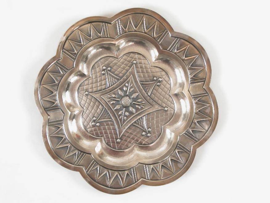 Apaļa dekoratīva metāla šķīvis ar reljefu zvaigžņveida un ziedu ornamentu.