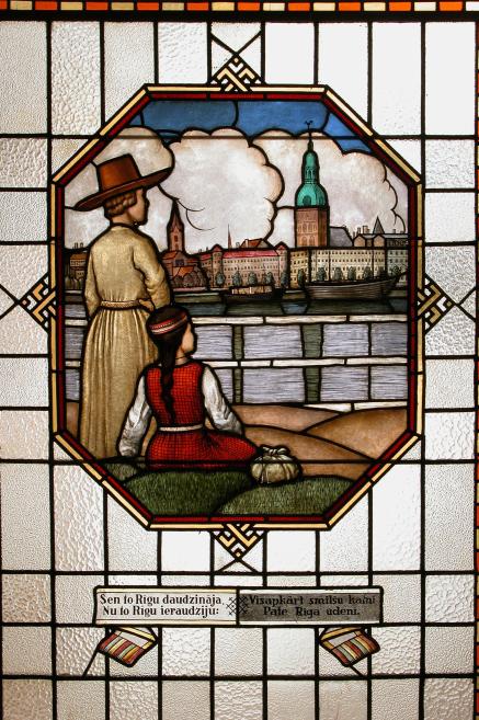 Krāsaina vitrāža ar sievieti un bērnu tradicionālos tērpos, kas skatās uz Rīgas panorāmu ar Sv. Pētera baznīcas torni un kuģiem Daugavā.