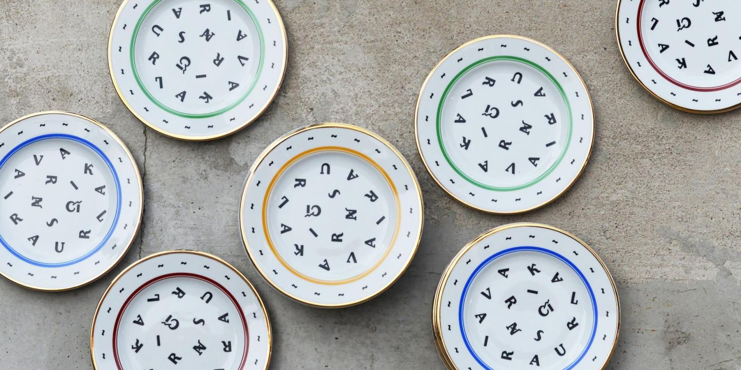 Six decorative white porcelain plates with colored rims and scattered black letters on a gray concrete surface.