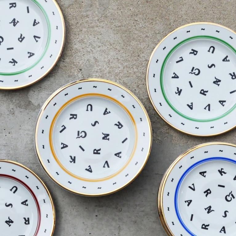 Six decorative white porcelain plates with colored rims and scattered black letters on a gray concrete surface.