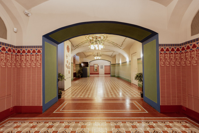 Muzeja interjera gaitenis ar dekoratīvām sienu flīzēm, ornamentētu arku, rakstainu flīžu grīdu un griestu lustru.