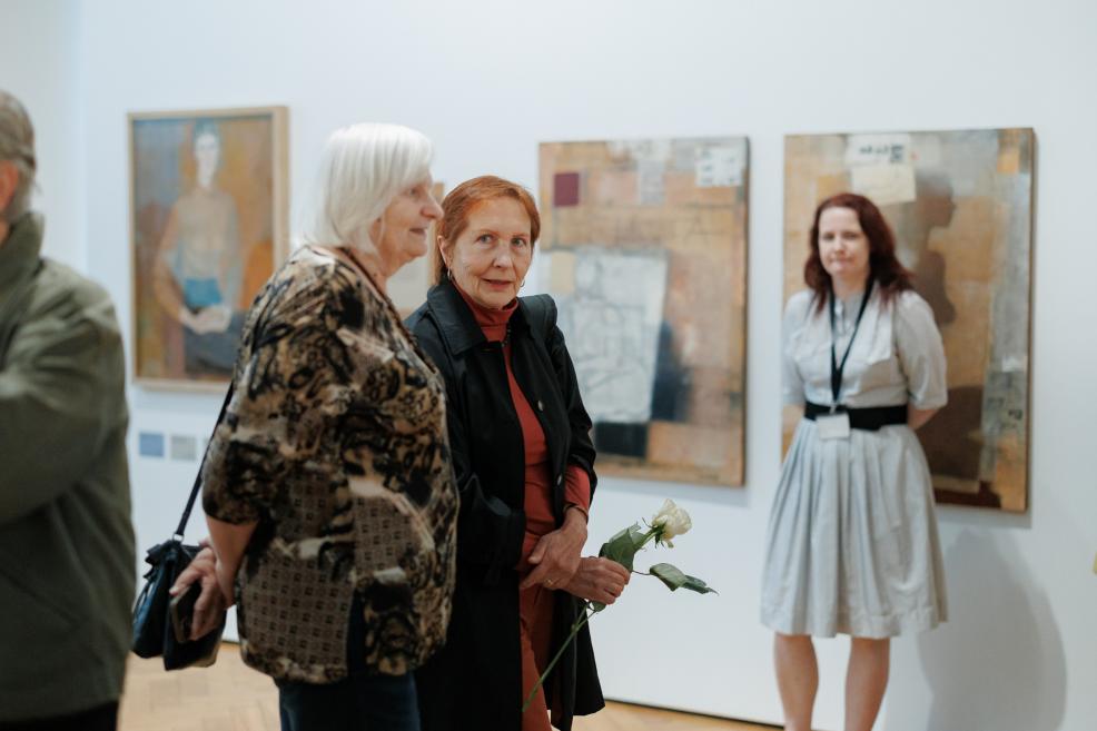 In an art gallery room, two older women stand, one holding a white rose, with several abstract paintings on the wall behind them and a museum visitor wearing a lanyard.