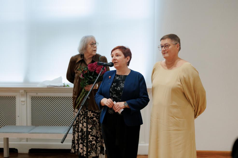 Three women stand by a microphone in an exhibition hall, one speaking while another behind her holds a bouquet of flowers.