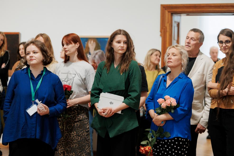 A group of people stands in a museum gallery holding flowers and folders, looking toward something outside the frame.