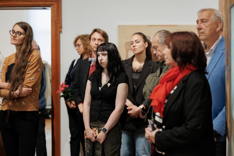 A small group of people stands in a museum gallery, listening or looking at something out of frame, against a white wall and wooden doorway.