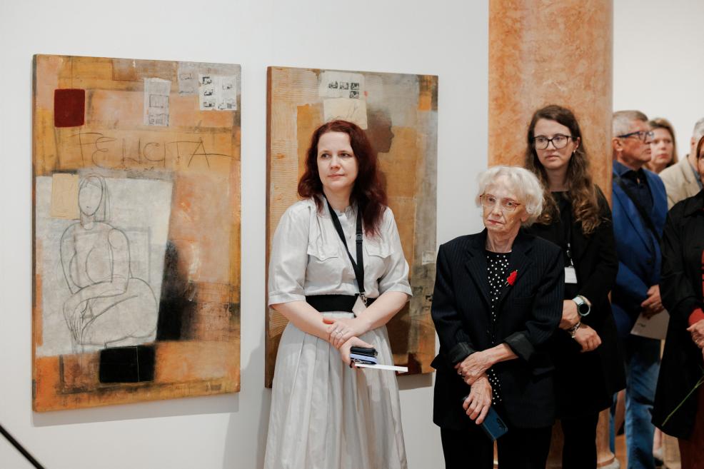 In a museum gallery, two large abstract paintings hang on the wall while several visitors stand in the foreground.