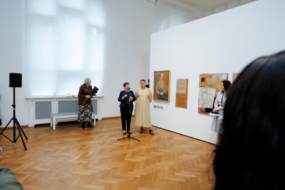 In a museum gallery, a woman speaks at a microphone beside exhibited paintings and an information panel, with several people standing in the background.