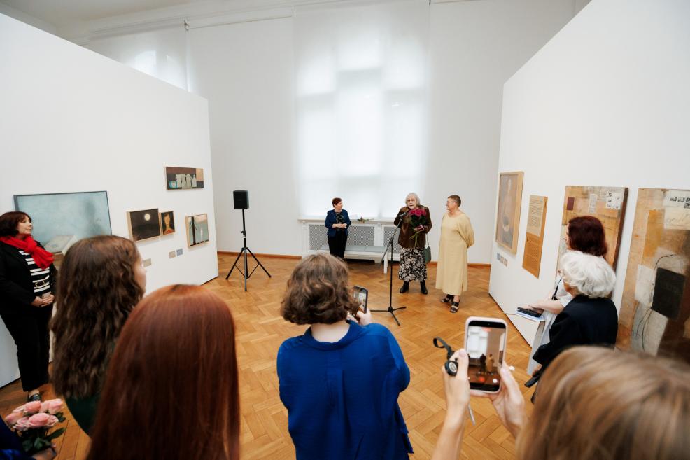 In a gallery room with parquet flooring and artworks mounted on the walls, a woman stands at a microphone holding flowers while visitors gather around her.