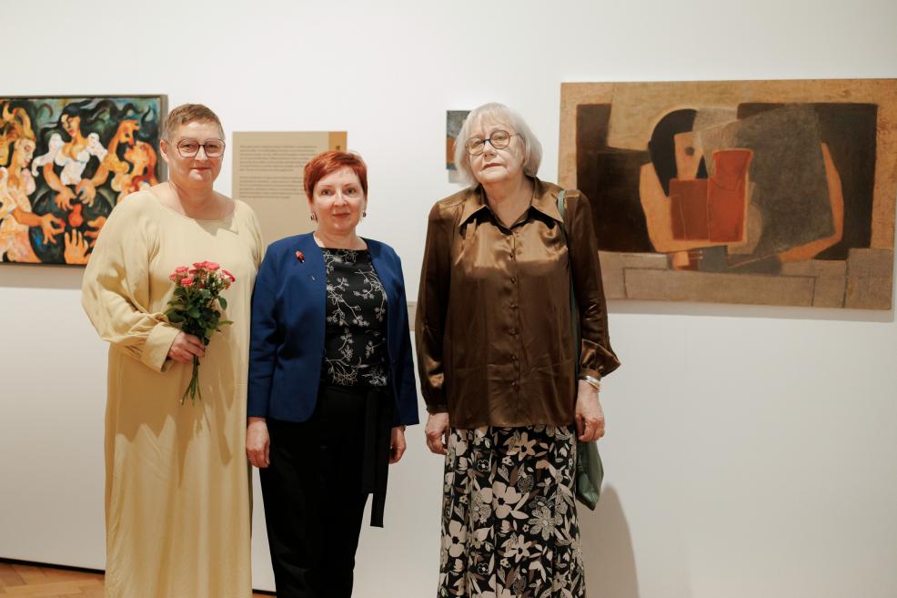 Three women stand in an art gallery beside a wall with paintings, one holding a bouquet of flowers.
