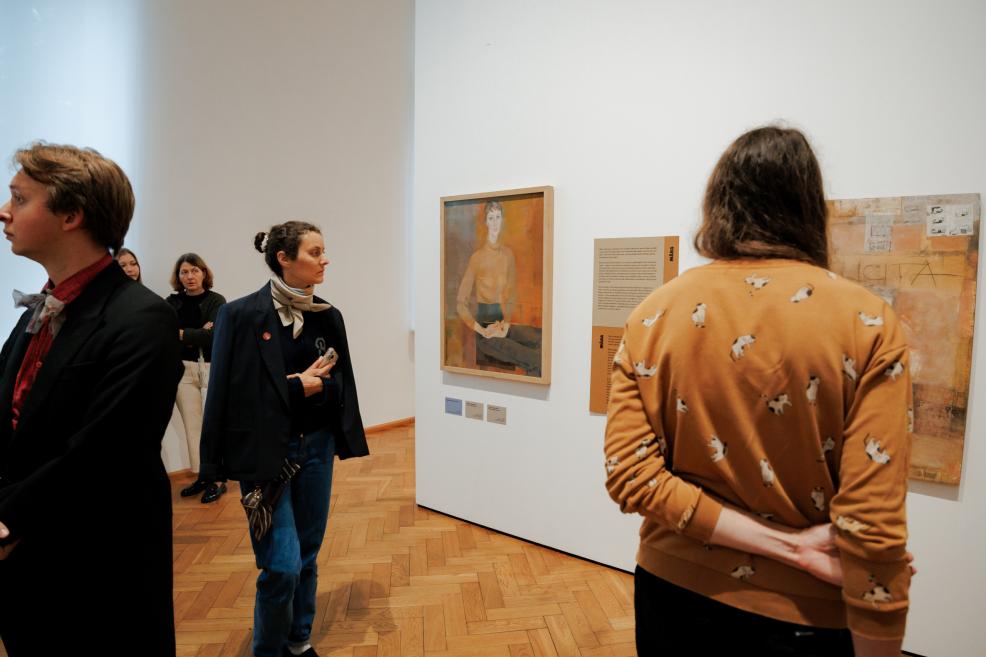In a museum gallery, visitors look at artworks displayed on a white wall, including a figurative painting and a larger work with text and collage elements.